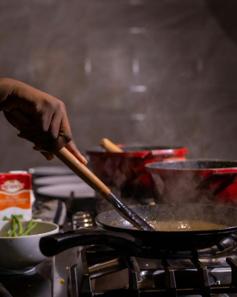 Chef's hand stirring a frying pan on a stove in a kitchen setting with dishes and food preparation ingredients.